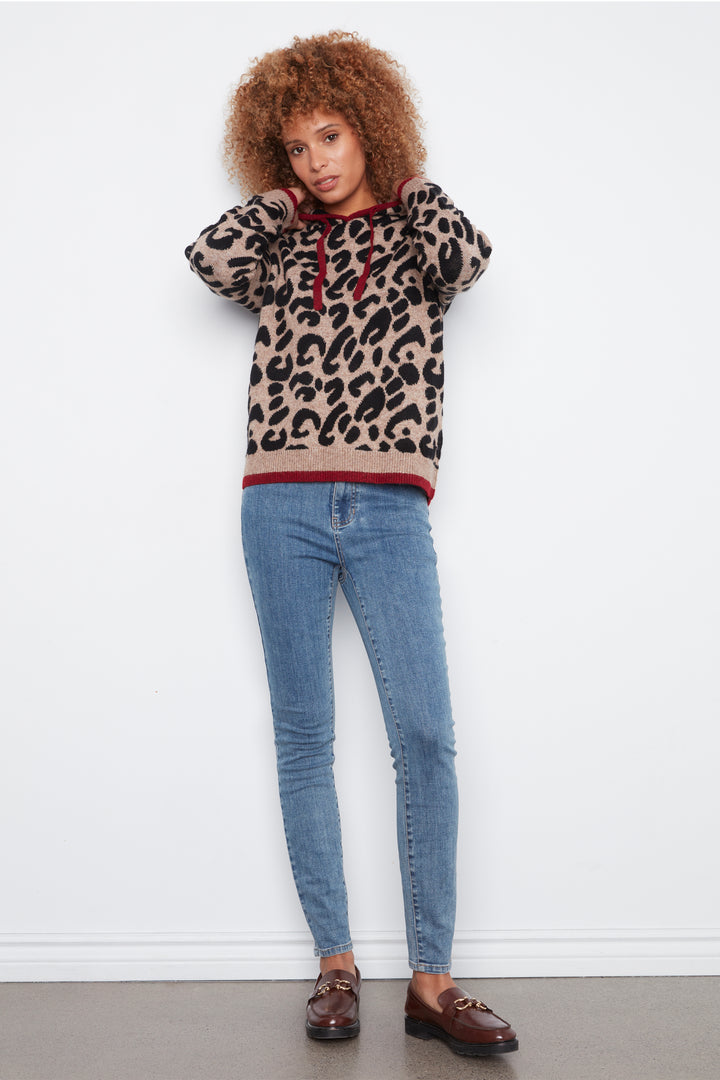 A woman with curly brown hair stands against a white wall wearing a beige and black leopard-print hoodie with red trim, blue skinny jeans, and brown loafers.