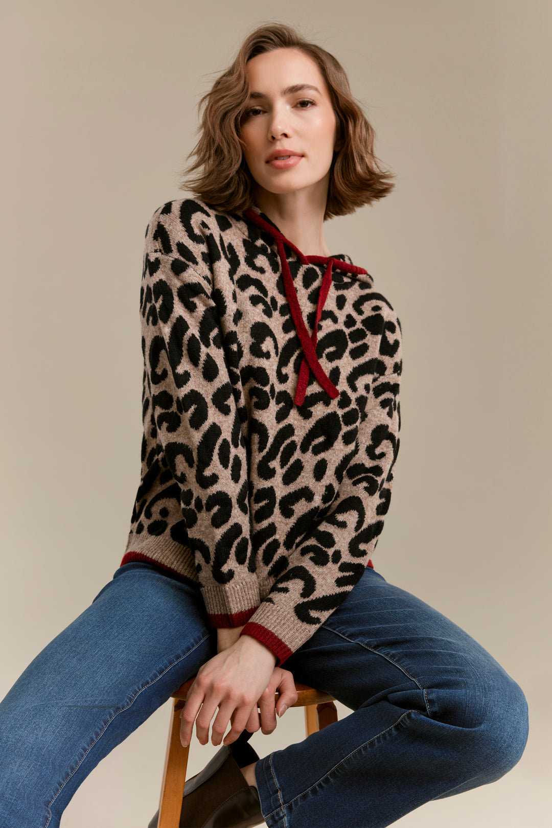 A woman seated on a wooden stool wearing a beige knit sweater with a black leopard print and dark red drawstrings and trim, paired with blue jeans, against a plain beige studio background.
