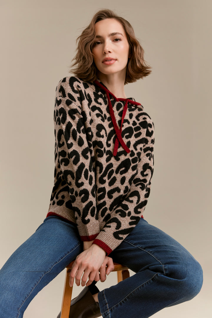 A woman seated on a wooden stool wearing a beige knit sweater with a black leopard print and dark red drawstrings and trim, paired with blue jeans, against a plain beige studio background.