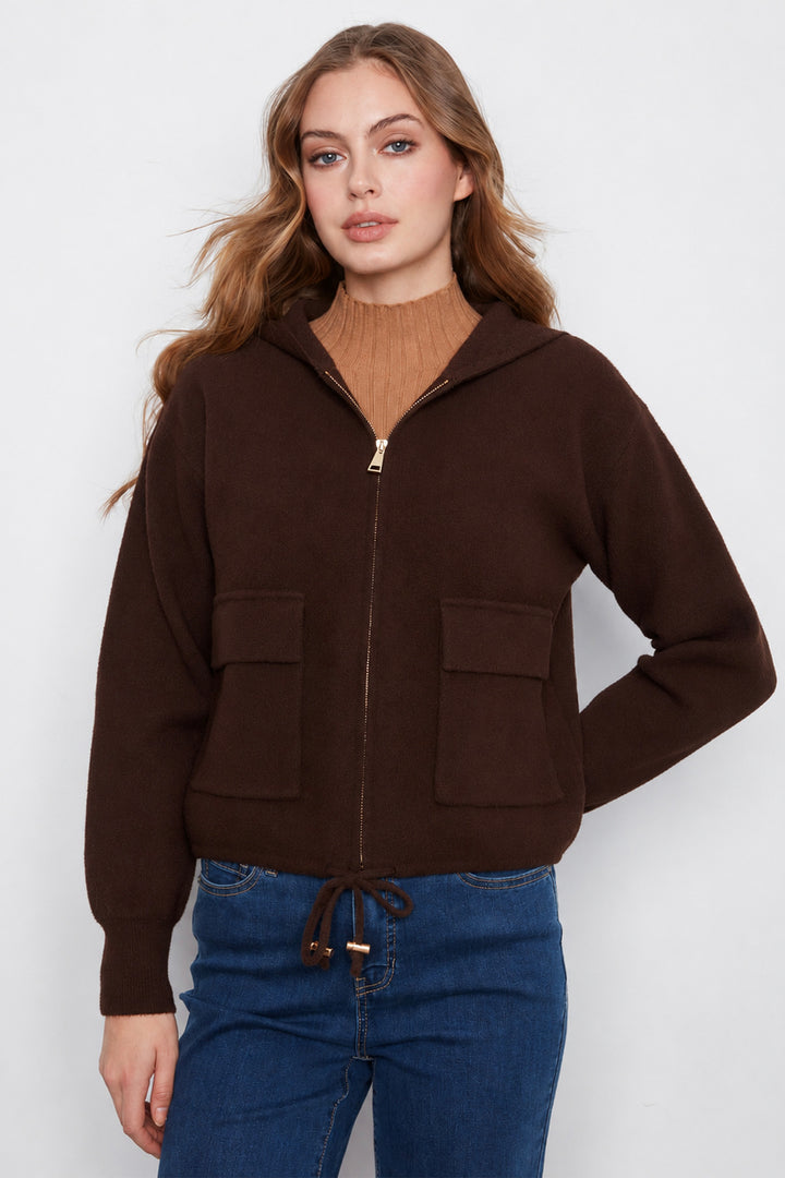 A woman wearing a dark brown zip-up hooded jacket with two front patch pockets, styled over a tan turtleneck and blue jeans, against a plain light gray studio background.