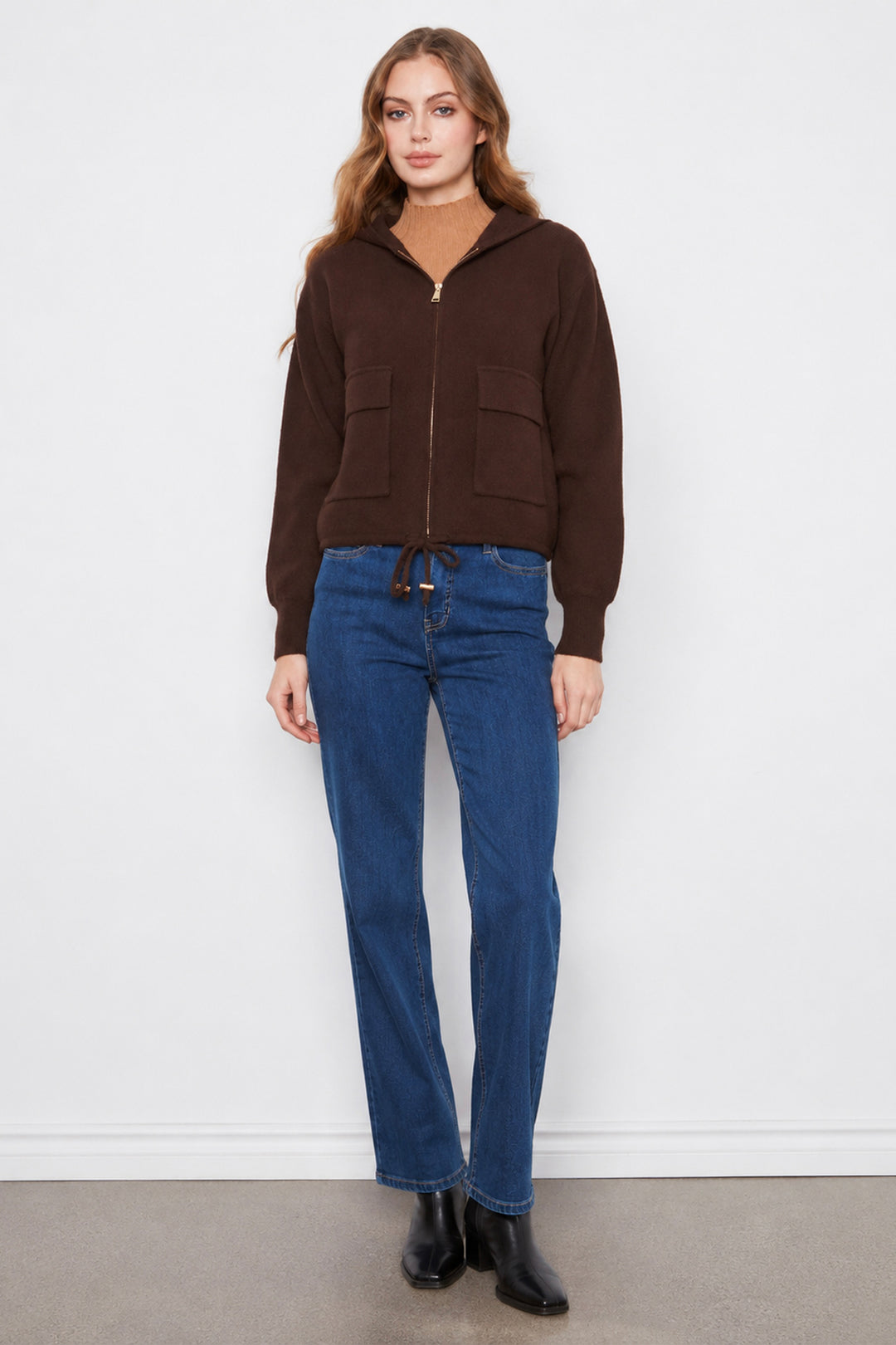 A woman modeling a dark brown cropped zip-up hoodie with front pockets, paired with blue jeans and black boots, standing against a plain white wall.