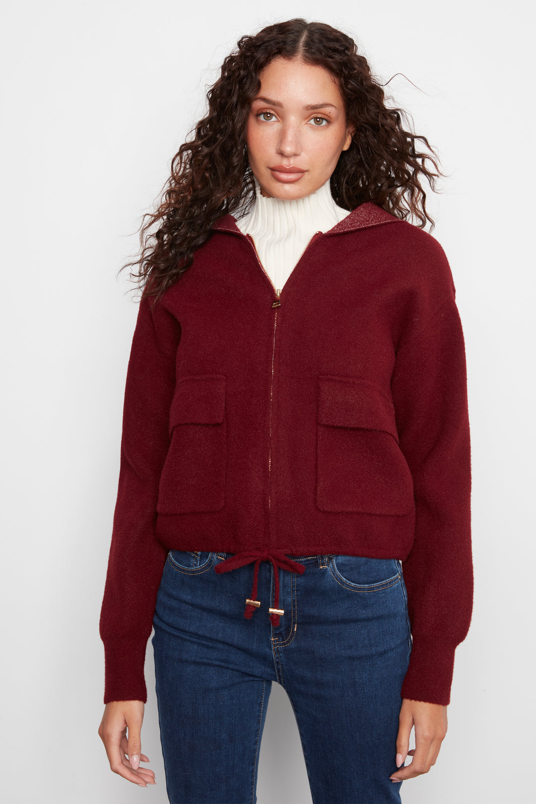 A woman wearing a cropped maroon zip-up hooded sweater with front patch pockets, styled over a white turtleneck and blue jeans, against a plain light gray background.