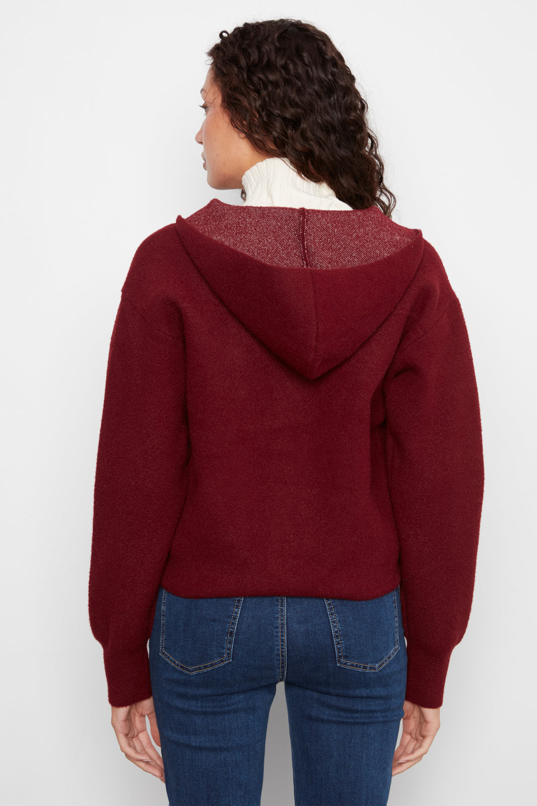 A woman shown from the back wearing a cropped maroon hooded sweater and blue jeans against a plain light gray studio background.