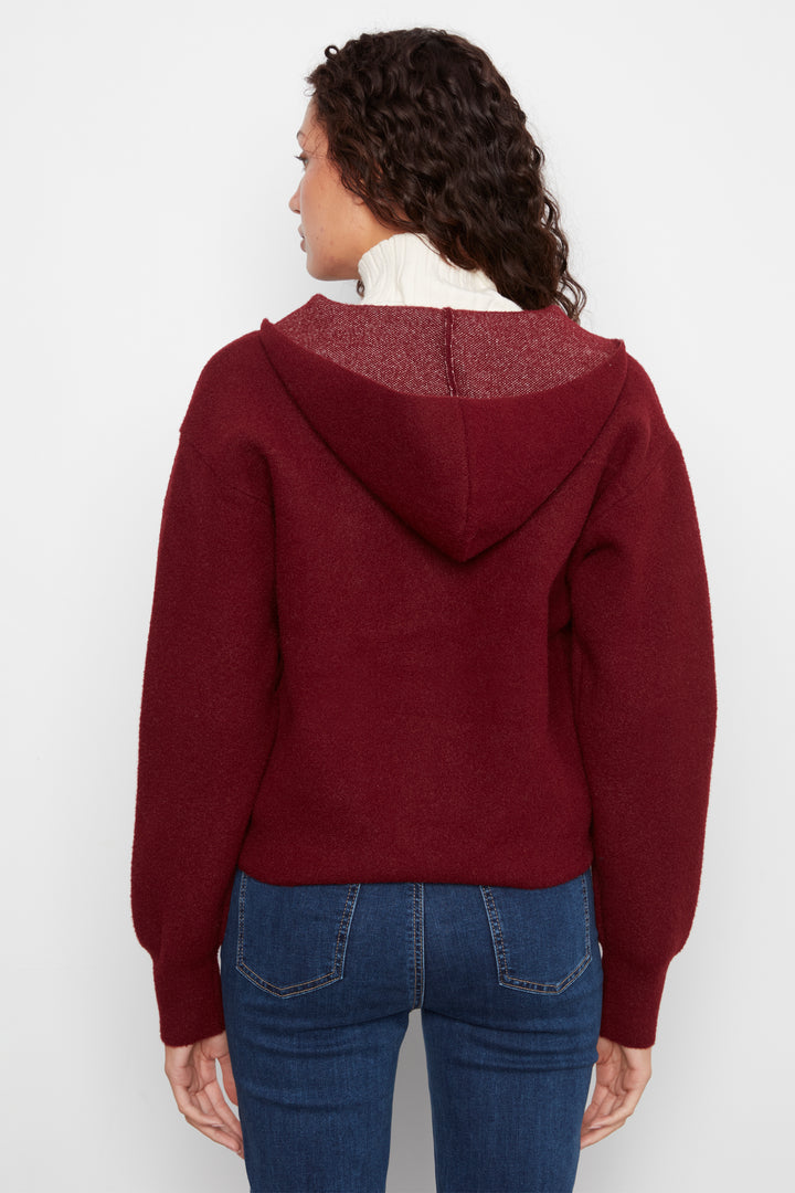 A woman shown from the back wearing a cropped maroon hooded sweater and blue jeans against a plain light gray studio background.