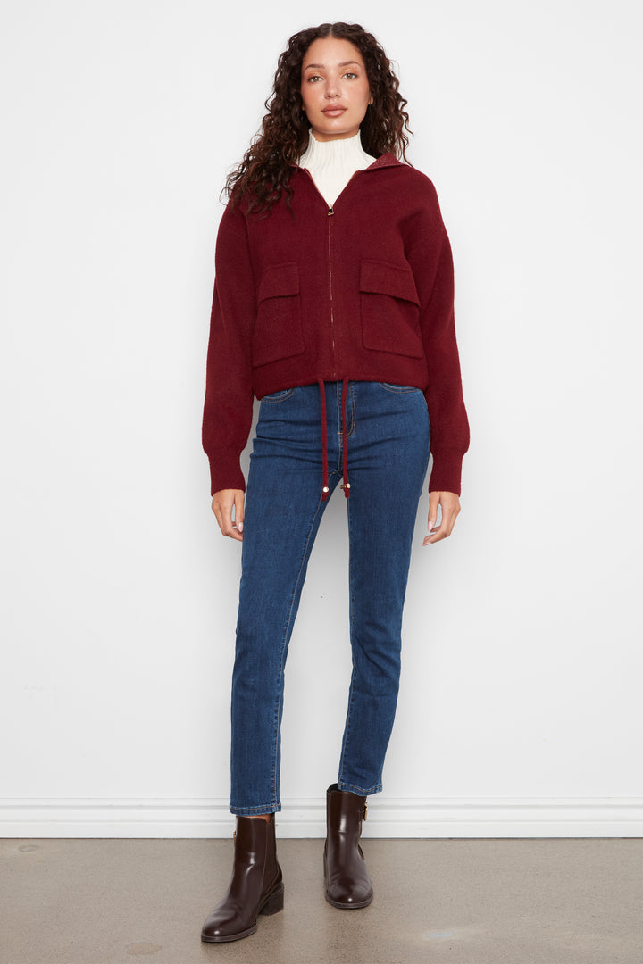 A model stands against a white wall wearing a burgundy zip-up knit cardigan with front patch pockets, blue jeans, and dark brown ankle boots.