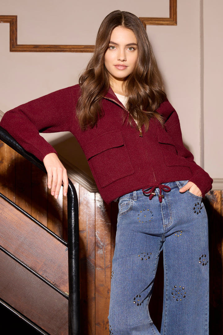 A woman models a burgundy half-zip knit sweater and distressed blue jeans while leaning against a wooden staircase railing in an indoor setting.