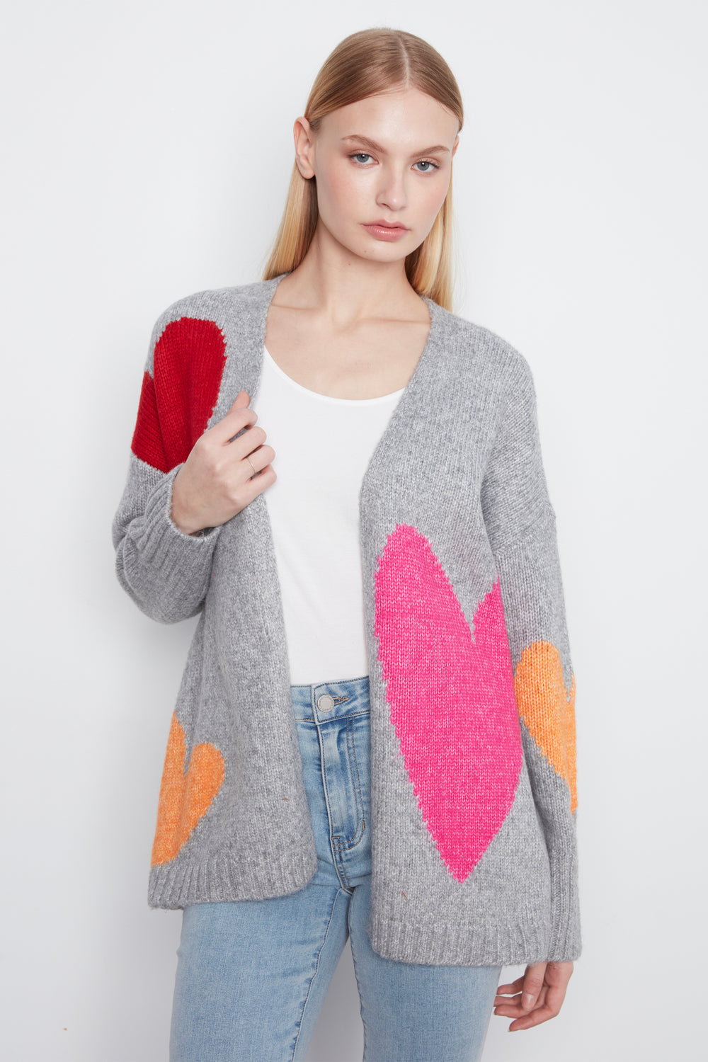 A woman modeling a gray knit cardigan with large colorful heart patches in red, pink, and orange, worn over a white top and light blue jeans against a plain light background.