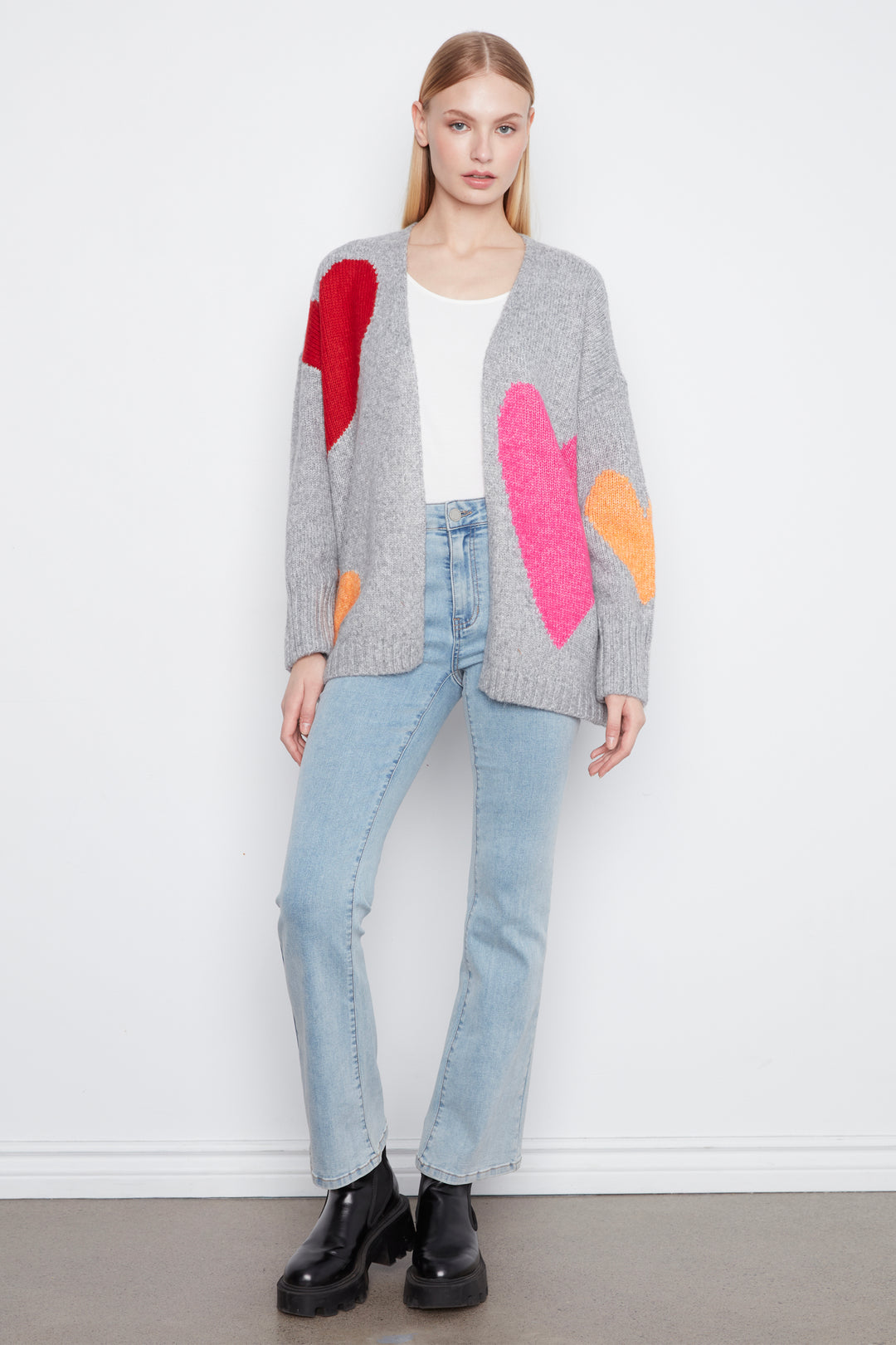 A model stands against a white wall wearing a light gray knit cardigan with large textured heart patches in red, pink, and orange, paired with light blue jeans and black boots.