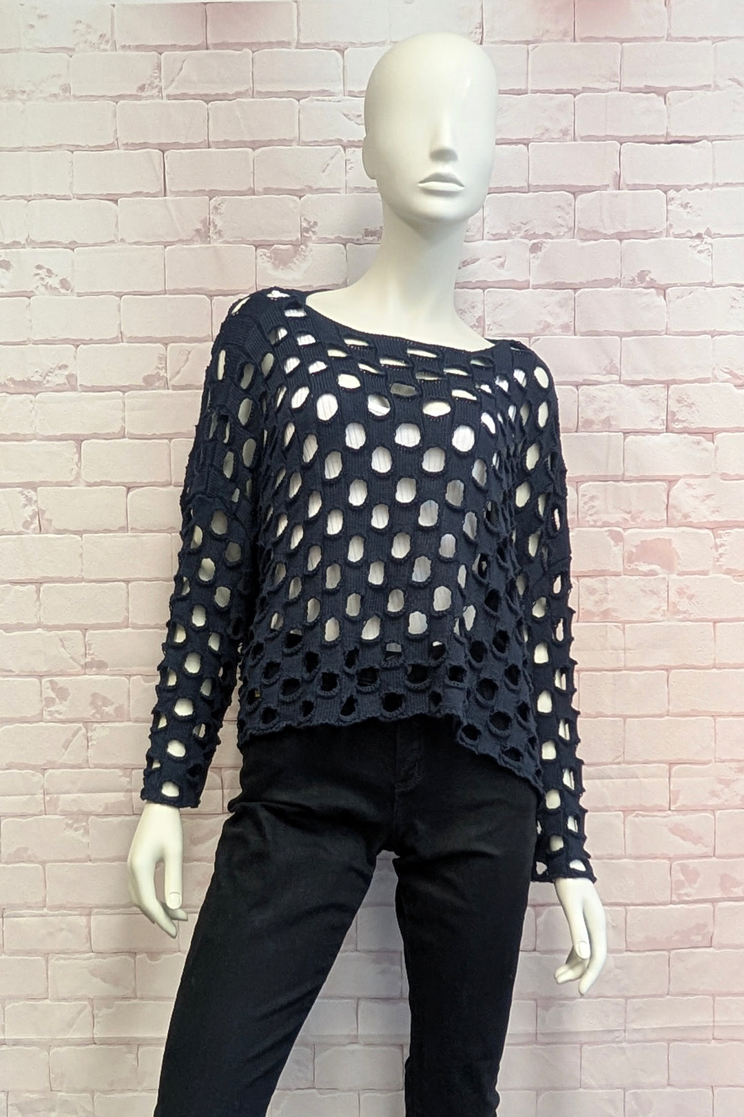 A mannequin wearing a black open-knit long-sleeve top with large circular cutouts and black pants, posed against a light pink brick wall.