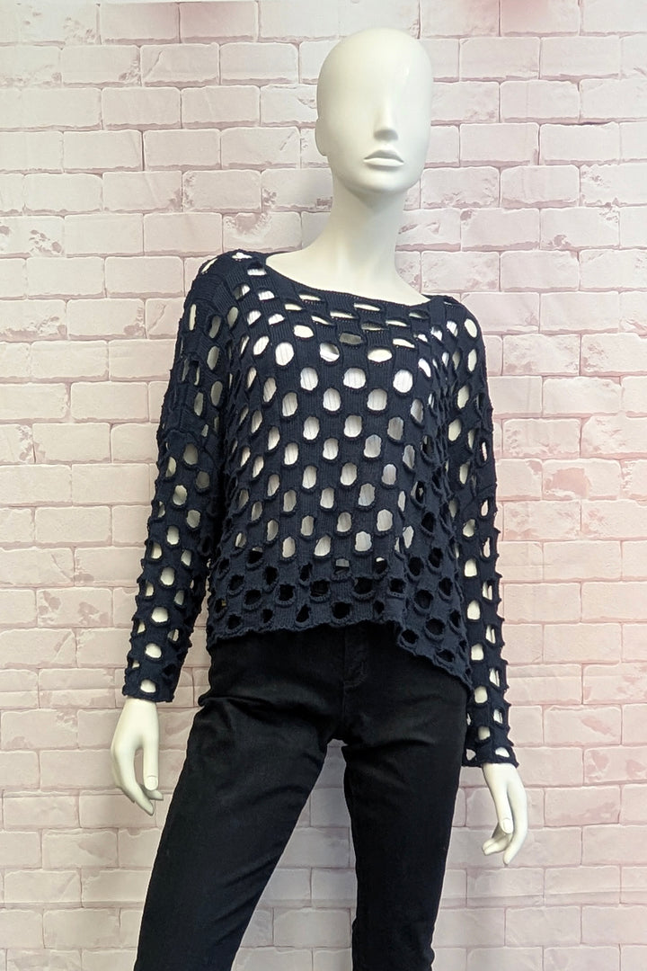 A mannequin wearing a black open-knit long-sleeve top with large circular cutouts and black pants, posed against a light pink brick wall.