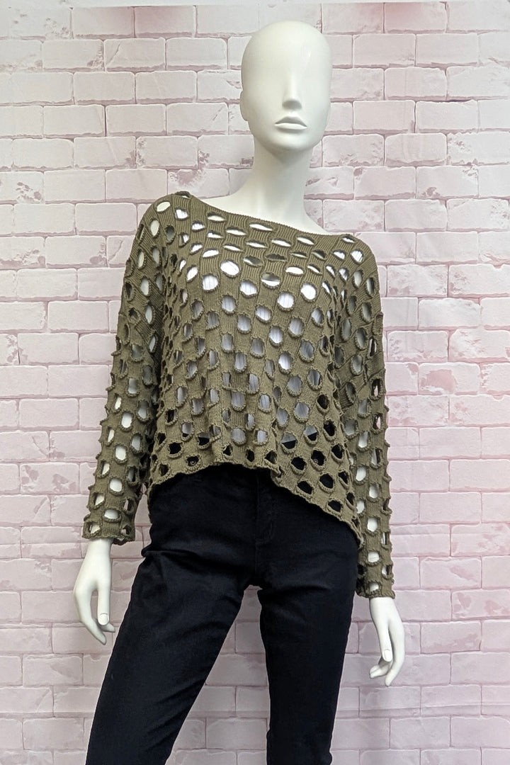 An olive green open-knit long-sleeve top displayed on a white mannequin, paired with black pants, set against a light pink brick wall background.