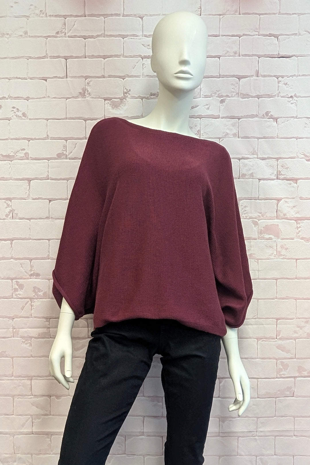 A mannequin wearing a loose, boat-neck knit top in deep burgundy with black pants, set against a light pink brick wall background.