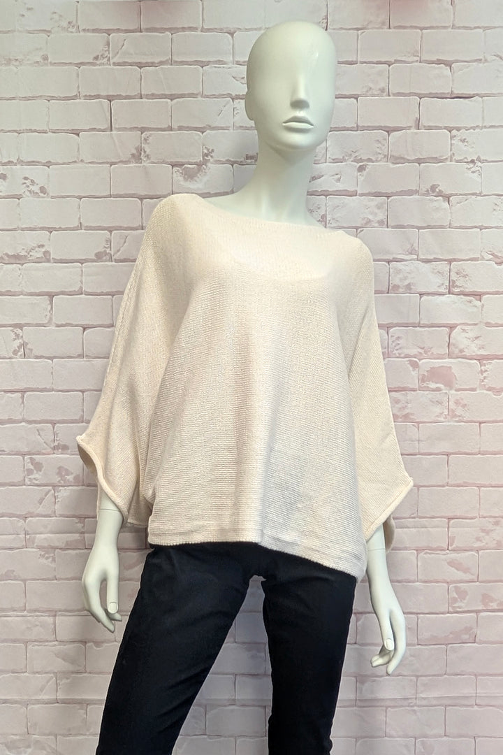 A mannequin wearing a loose, short-sleeve knit top in a light cream or off-white color, paired with black pants, set against a light pink brick wall background.