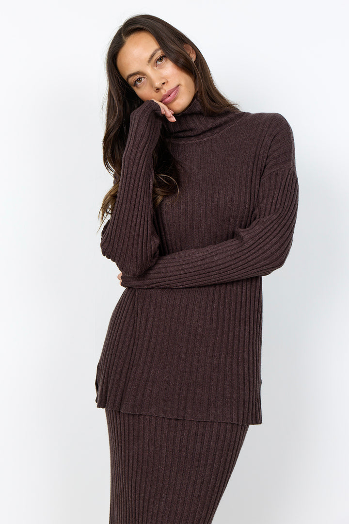 A woman wearing a dark brown ribbed knit turtleneck sweater and matching ribbed skirt against a plain white studio background.