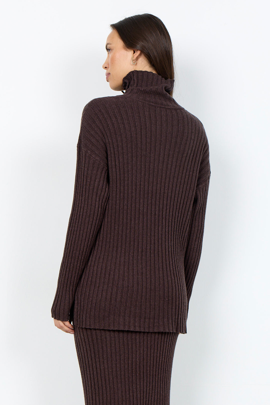 A woman shown from the back wearing a dark brown ribbed turtleneck sweater and matching ribbed skirt against a plain light gray studio background.