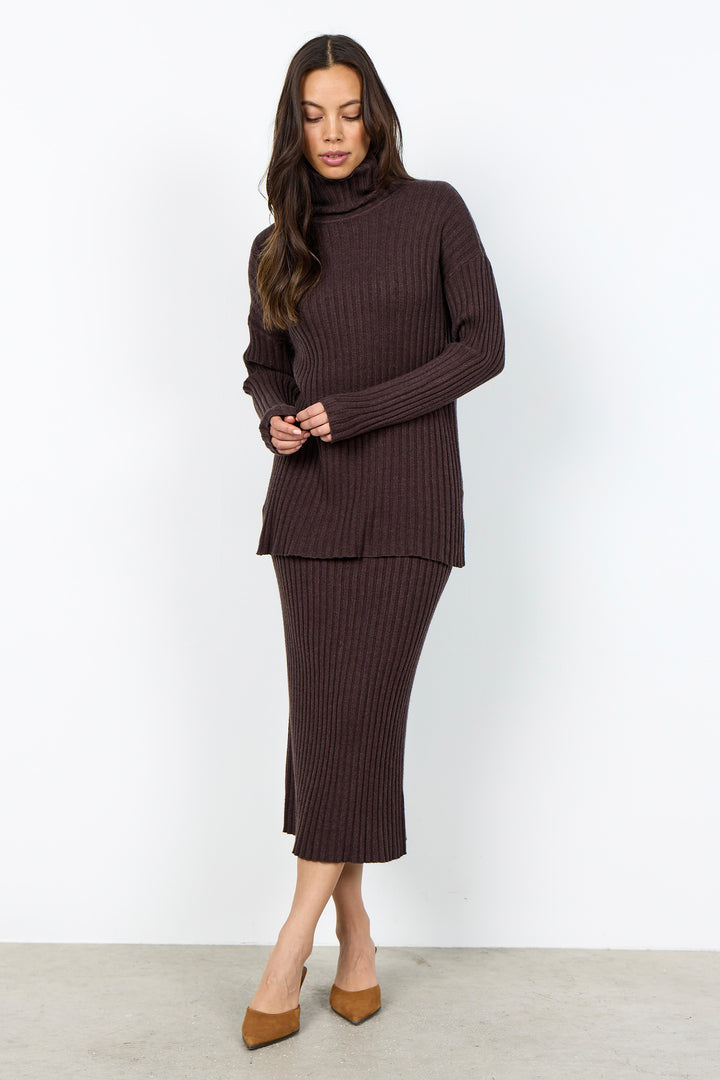 A woman modeling a dark brown ribbed knit turtleneck sweater and matching midi skirt set, paired with tan pointed-toe heels, against a plain white studio background.