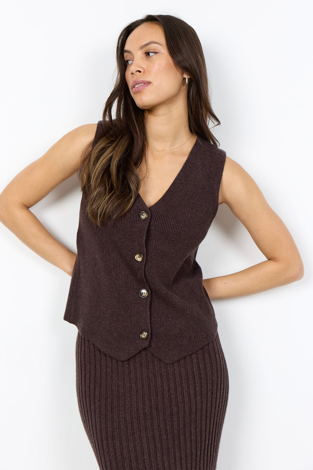 A woman modeling a sleeveless ribbed knit vest in dark brown with three front buttons, paired with a matching ribbed knit skirt, against a plain white studio background.