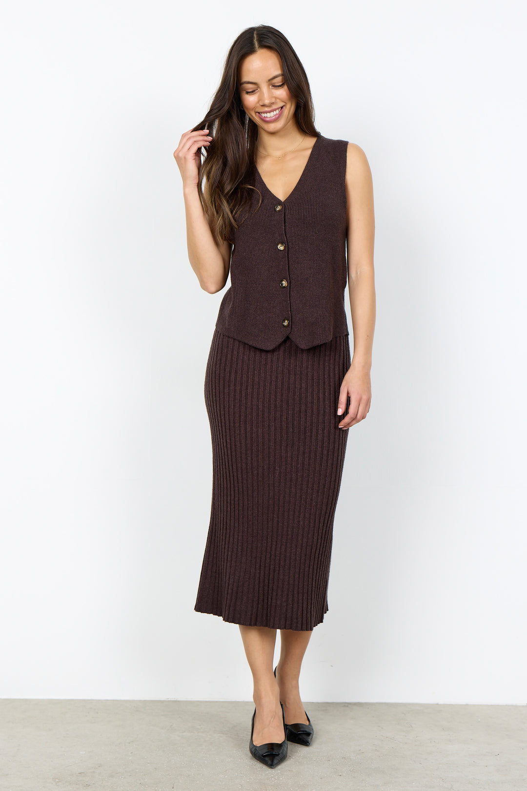 A woman modeling a dark brown ribbed knit vest and midi skirt set against a plain white studio background.