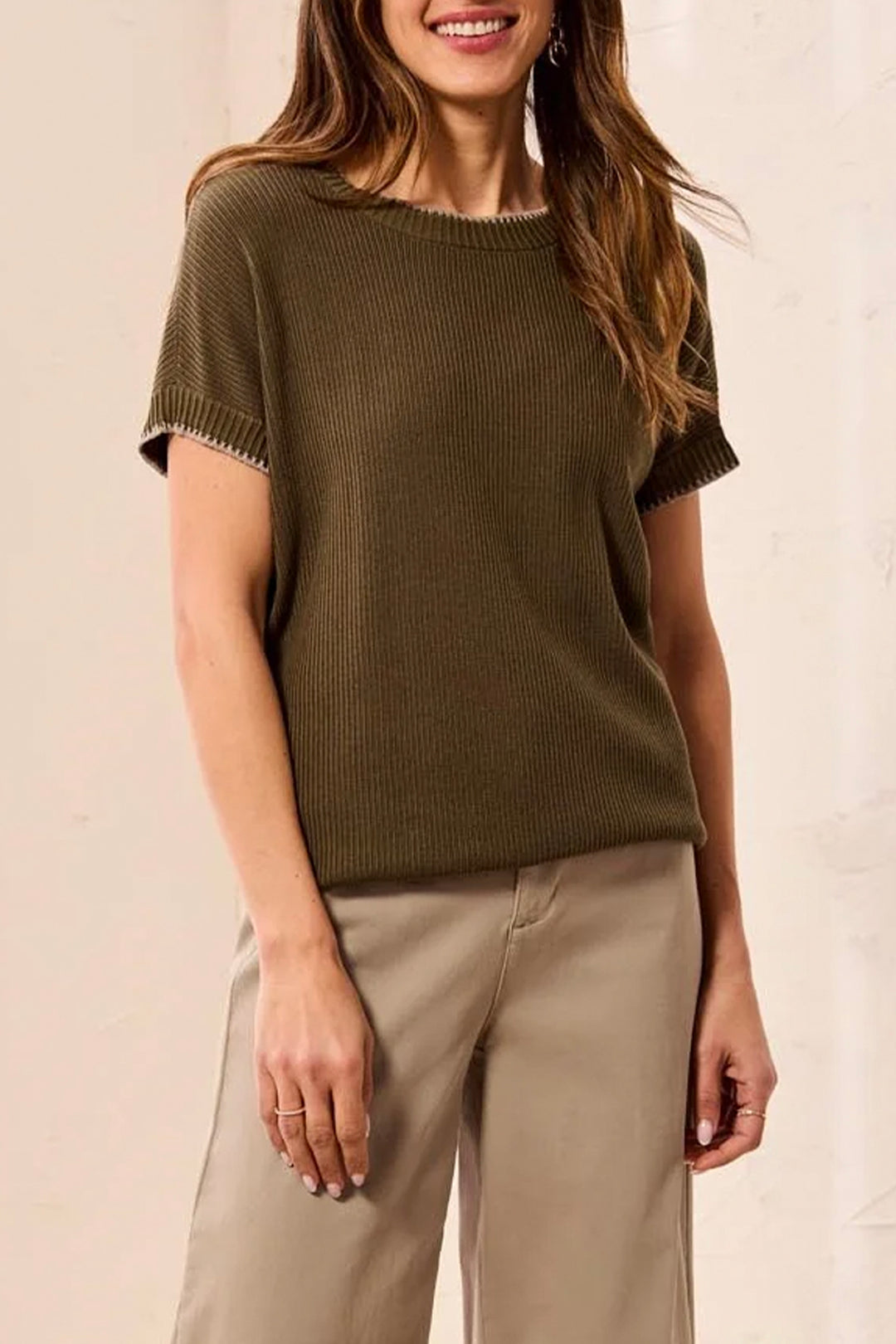 A woman wearing a short-sleeve ribbed knit top in olive green with beige wide-leg pants, standing against a light beige wall.