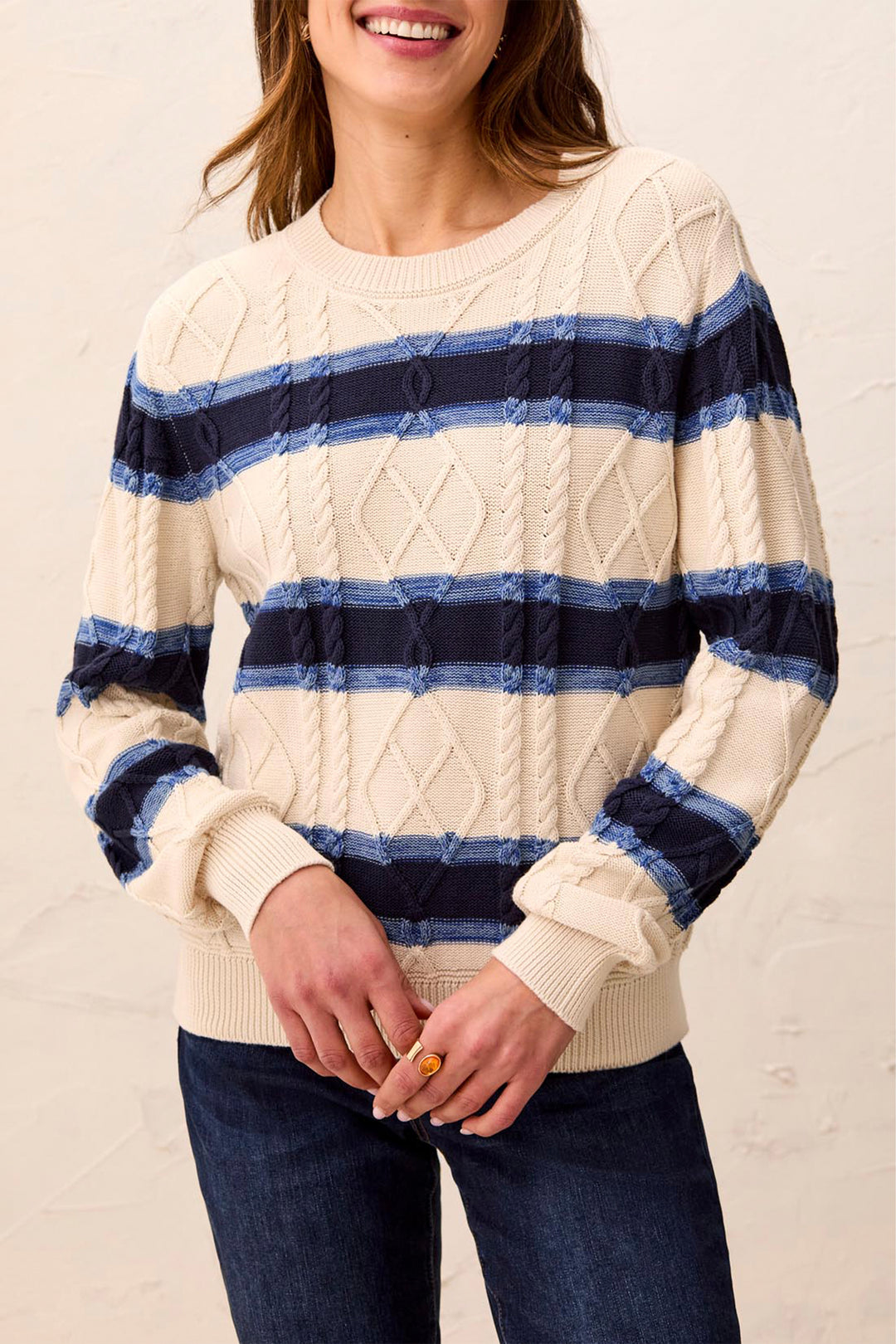 A woman wearing a cream cable-knit sweater with horizontal navy and medium blue stripes, paired with dark blue jeans, against a light beige textured wall.