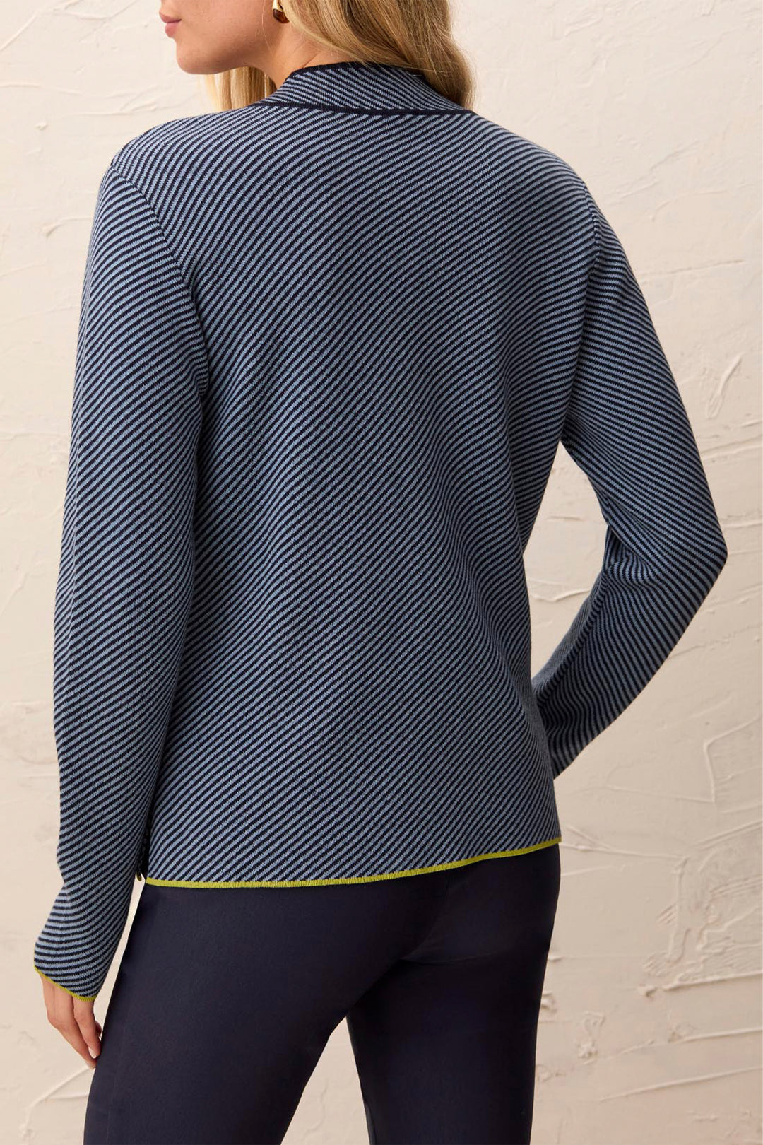 A woman shown from the back wearing a navy blue long-sleeve knit top with thin diagonal white stripes and a thin yellow-green hem trim, paired with dark leggings against a textured off-white wall.