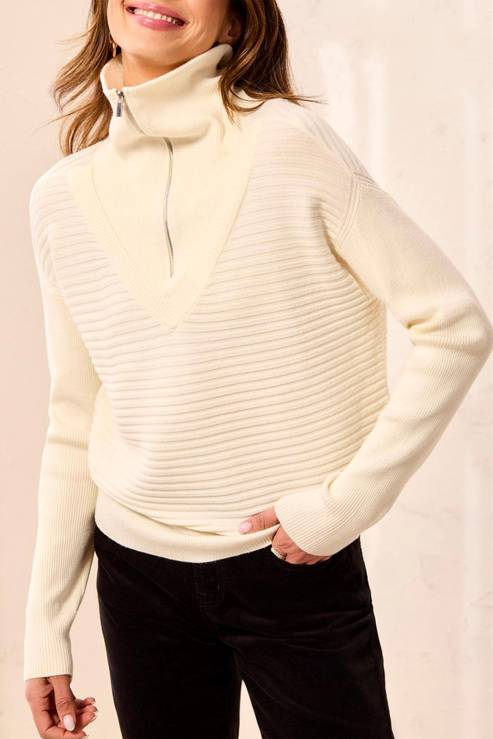 A woman wearing a cream ribbed knit mock neck sweater with a half-zip collar, paired with black pants, against a light beige background.