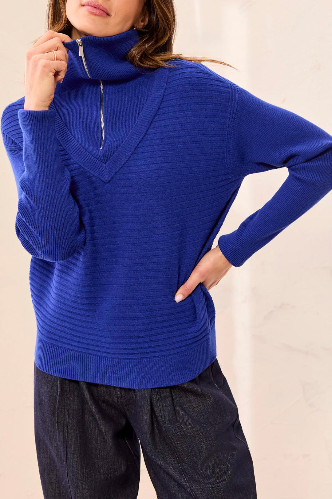 A woman wearing a bright royal blue ribbed knit sweater with a high folded collar and a short front zipper, paired with dark pants.