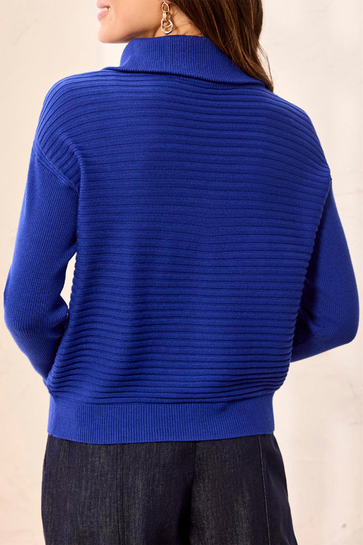 A woman wearing a royal blue ribbed knit mock neck sweater, shown from the back against a light beige background.