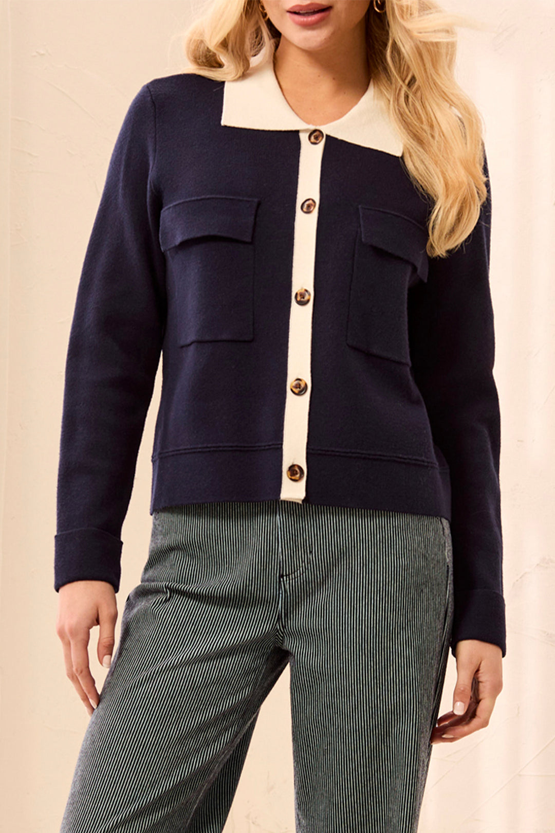 A woman wearing a navy blue cropped cardigan with a cream-colored collar and button placket, paired with green pinstriped trousers, against a neutral beige background.