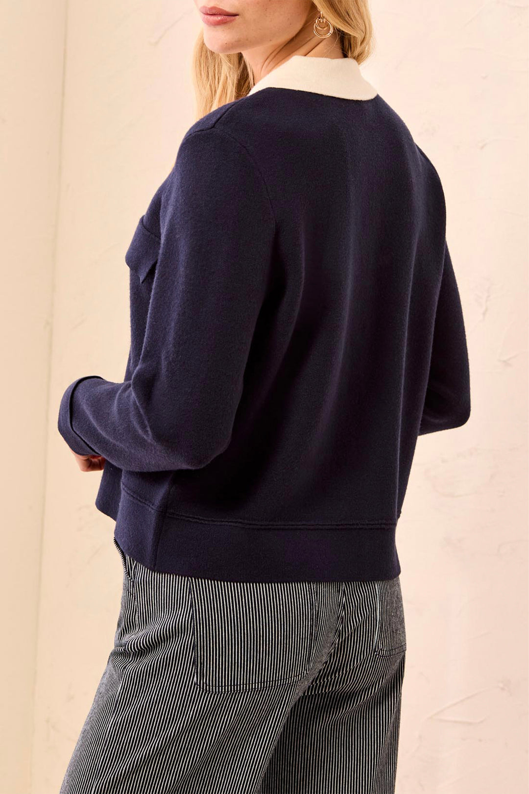 A woman shown from the back wearing a navy blue knit sweater with a contrasting white crew neck collar, paired with gray pinstriped trousers, against a light beige textured wall.
