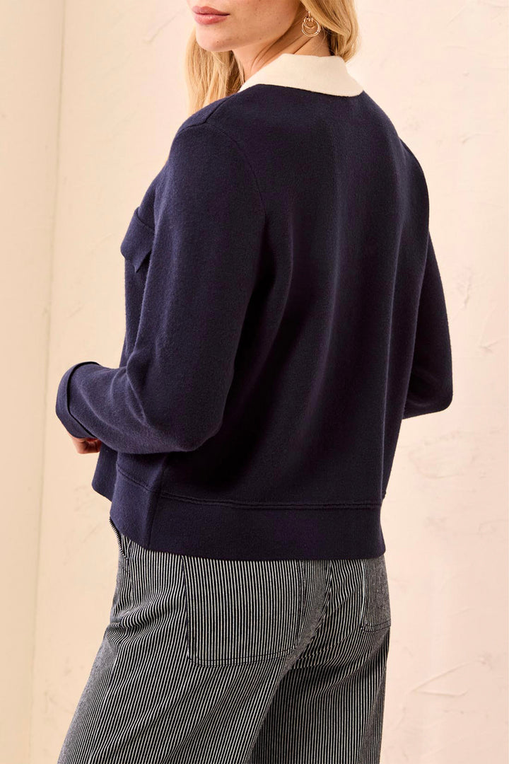A woman shown from the back wearing a navy blue knit sweater with a contrasting white crew neck collar, paired with gray pinstriped trousers, against a light beige textured wall.