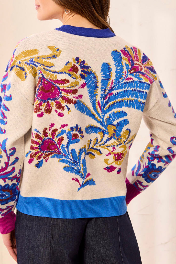 A close-up back view of a person wearing a cream knit sweater with large multicolored floral and leaf patterns in blue, magenta, and gold, plus blue ribbed collar, hem, and cuffs.