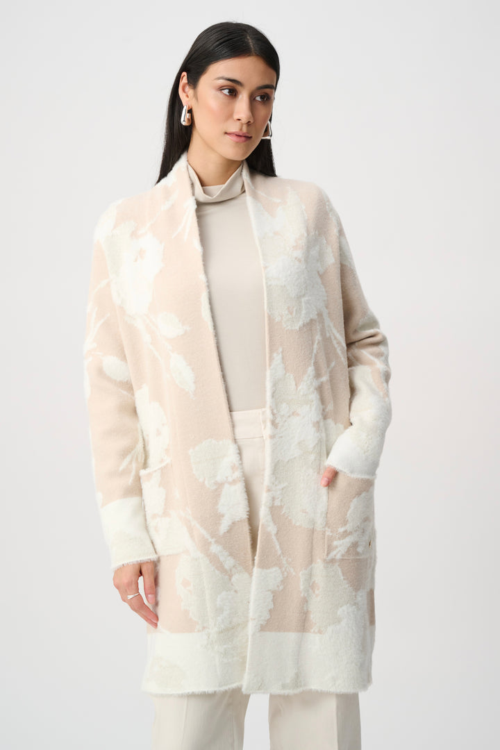 A woman modeling a long open-front cardigan in a soft blush beige and white floral pattern, worn over a matching beige turtleneck and cream pants against a light gray studio background.