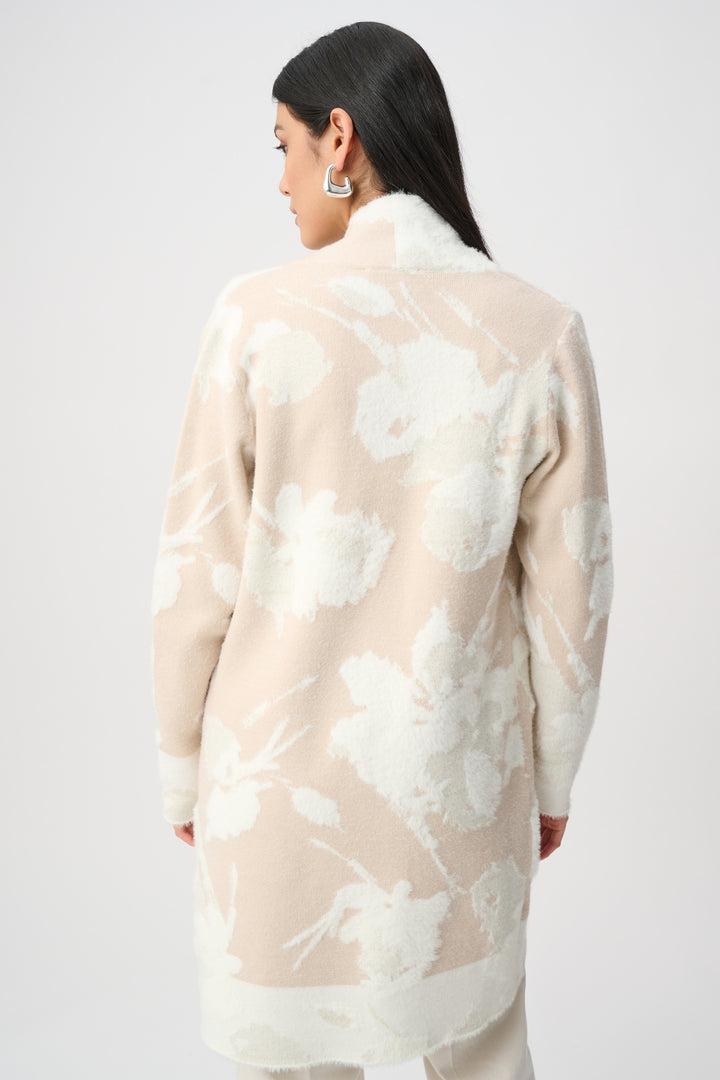 A woman modeling a long open-front cardigan in a soft blush beige and white floral pattern, worn over a matching beige turtleneck and cream pants against a light gray studio background.