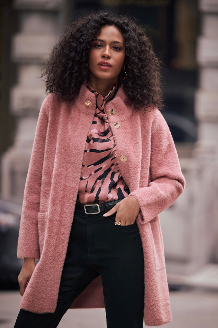 A woman wearing a dusty pink textured bouclé coat over a pink and black patterned blouse, paired with black trousers, standing outdoors against a blurred city background.