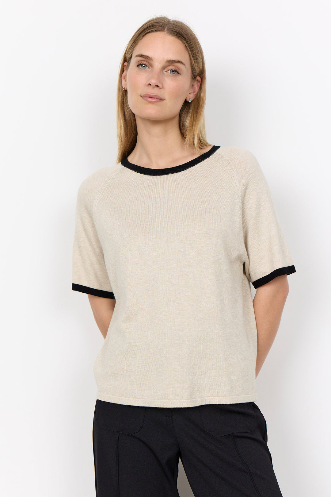 A woman modeling a short-sleeve knit top in warm off-white with black trim at the crew neck and sleeve cuffs, paired with black pants, against a plain white studio background.