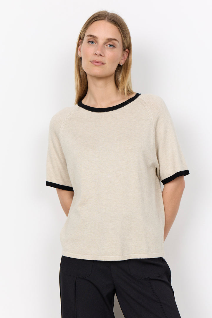 A woman modeling a short-sleeve knit top in warm off-white with black trim at the crew neck and sleeve cuffs, paired with black pants, against a plain white studio background.