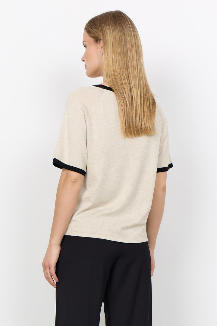 A woman shown from the back wearing a short-sleeve knit top in warm off-white with black trim at the collar and sleeve cuffs, paired with black pants, against a plain light gray studio background.