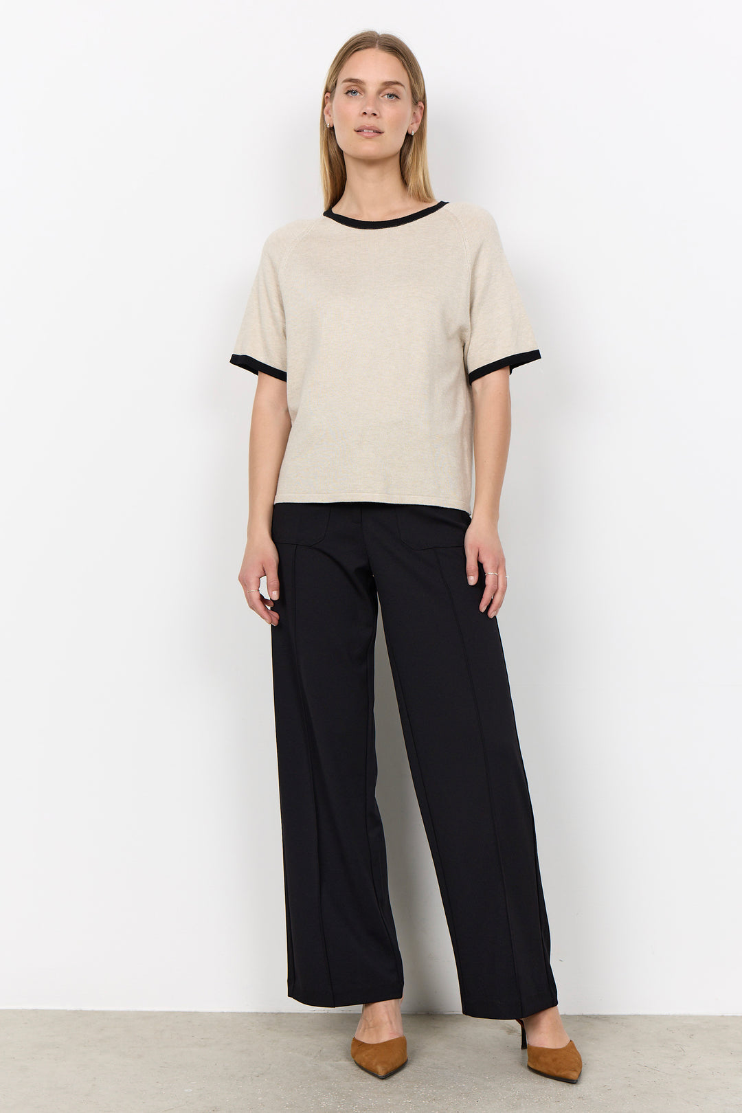 A woman modeling a short-sleeve knit top in warm off-white with black trim at the crew neck and sleeve cuffs, paired with black wide-leg trousers and tan pointed flats, against a plain white studio wall.