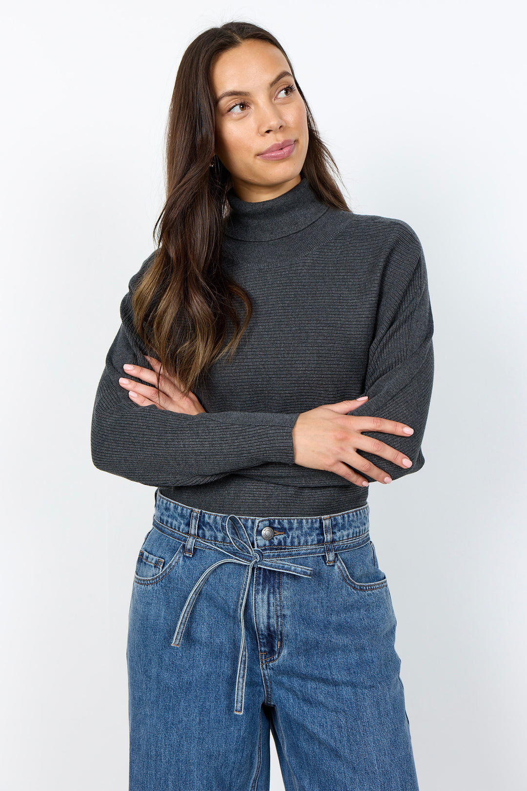 A woman wearing a dark charcoal gray ribbed mock neck sweater and blue jeans, standing against a plain white studio background with her arms crossed.