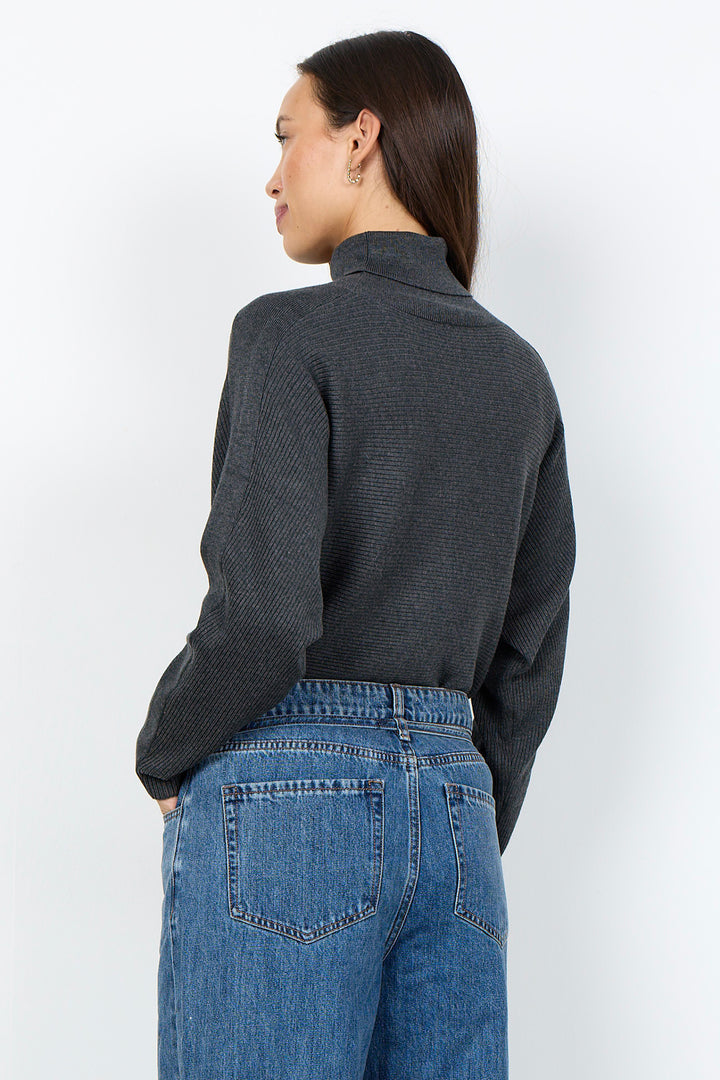 A woman shown from the back wearing a dark charcoal gray ribbed knit mock neck sweater and blue jeans, standing against a plain white studio background.