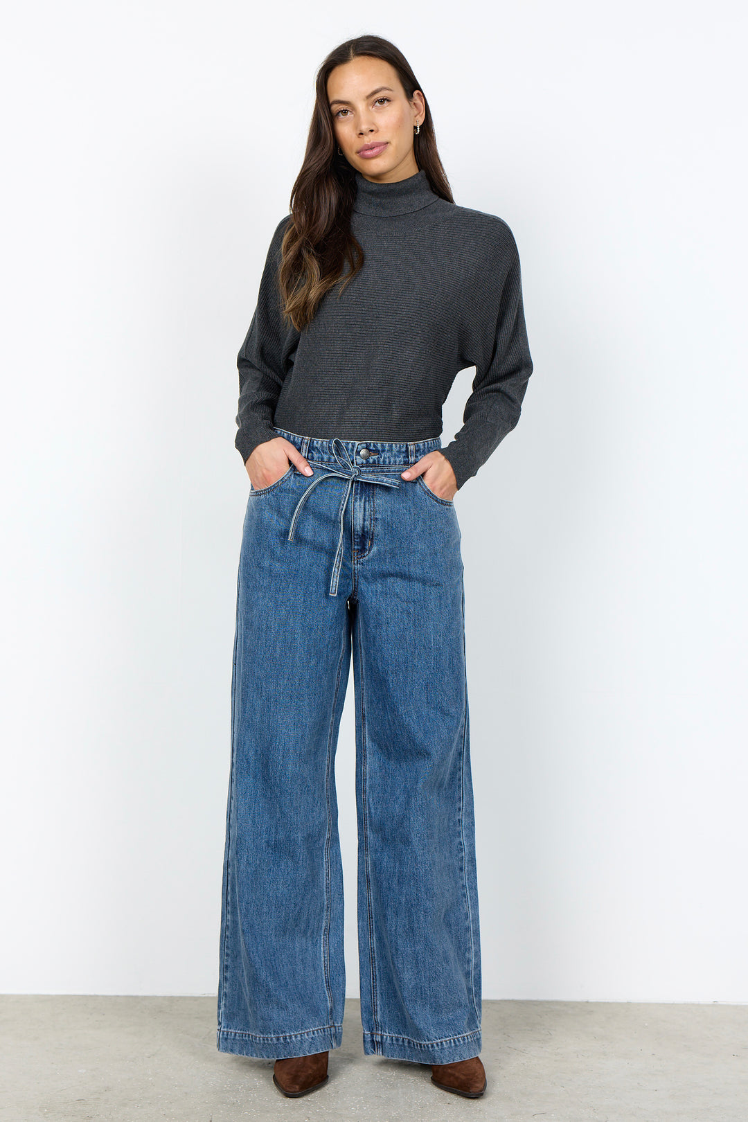 A woman modeling a dark charcoal gray mock neck knit top and high-waisted, wide-leg blue denim jeans with a drawstring waist, standing against a plain white studio background.
