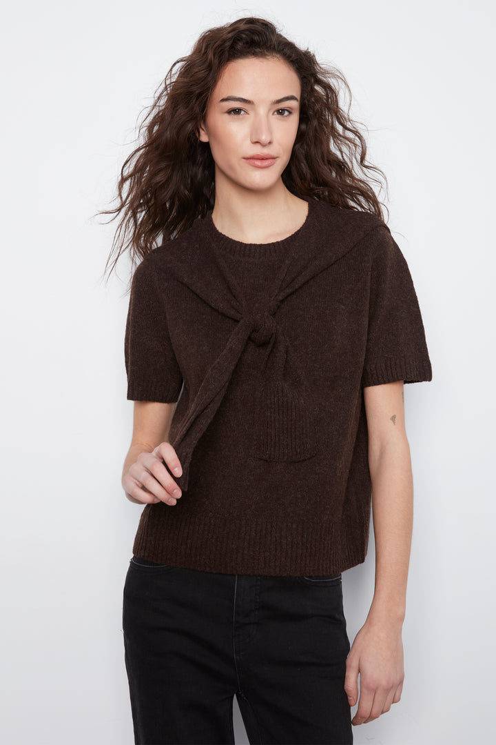 A woman modeling a short-sleeve knit top in dark brown with a twisted front detail, paired with black pants, against a plain light gray background.