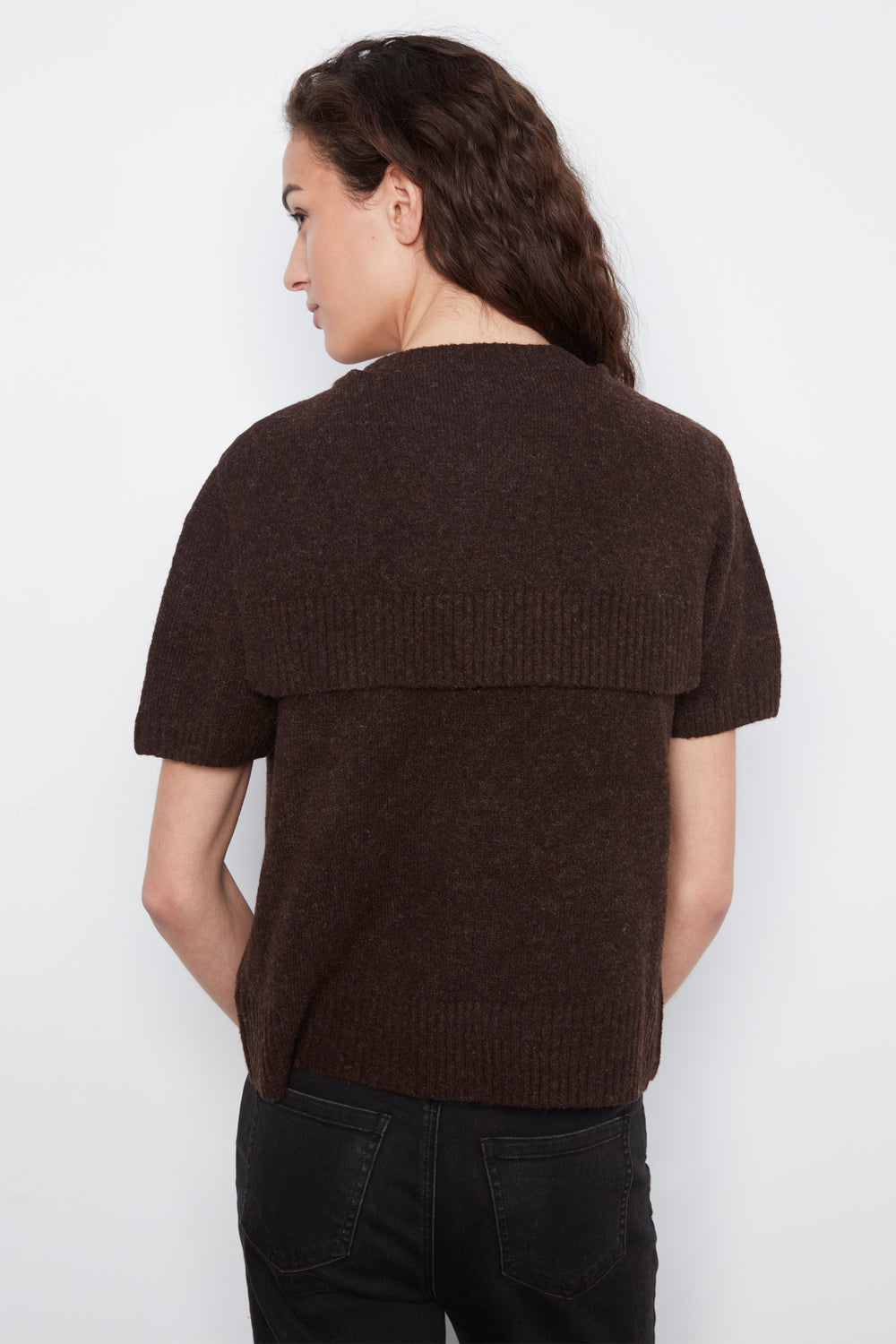 A woman shown from the back wearing a dark brown short-sleeve knit sweater with a horizontal ribbed band across the upper back, paired with black jeans, against a plain light gray background.