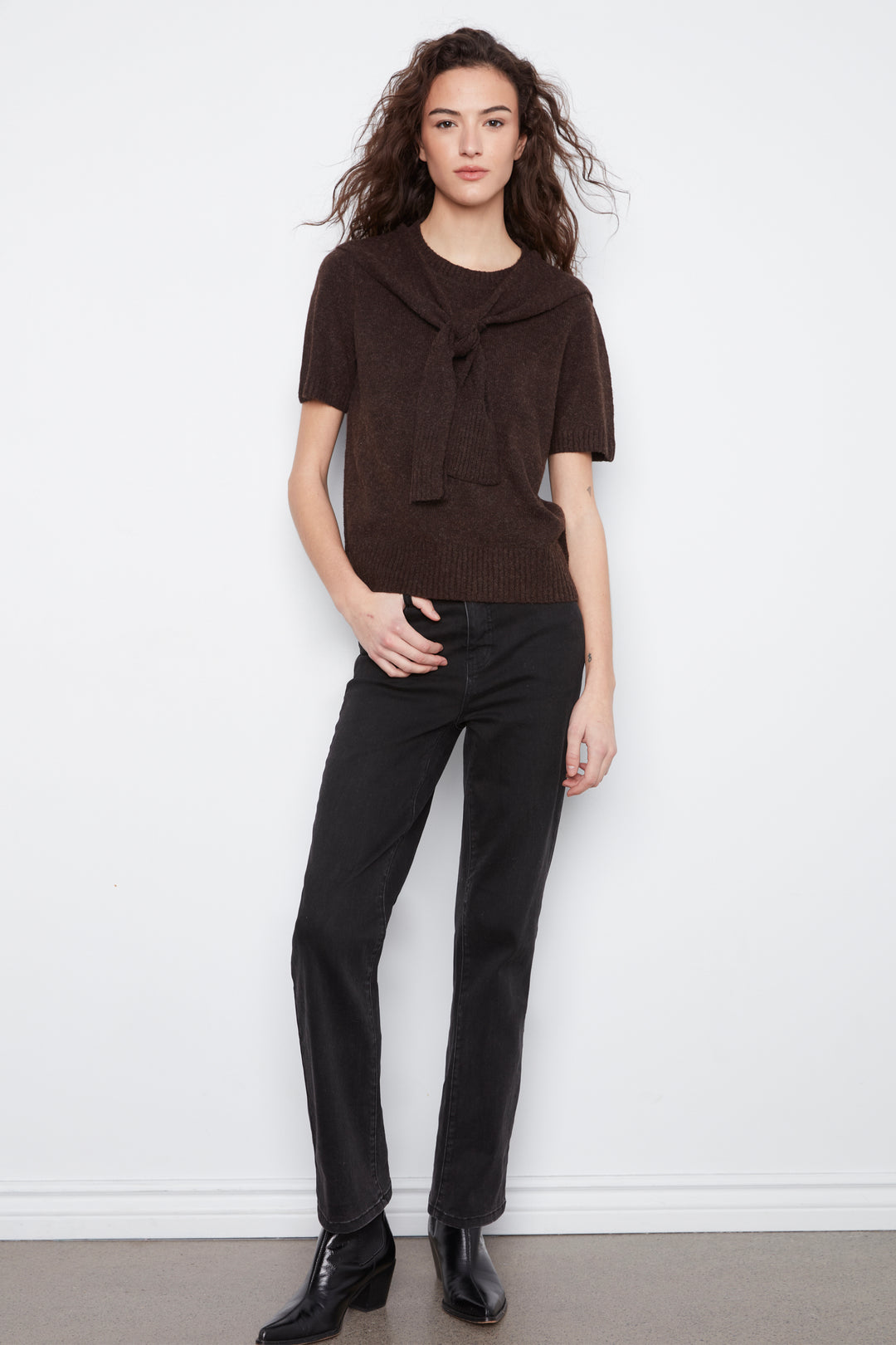 A woman modeling a dark brown short-sleeve knit top with a large front tie detail, paired with black pants and black ankle boots, standing against a white wall.