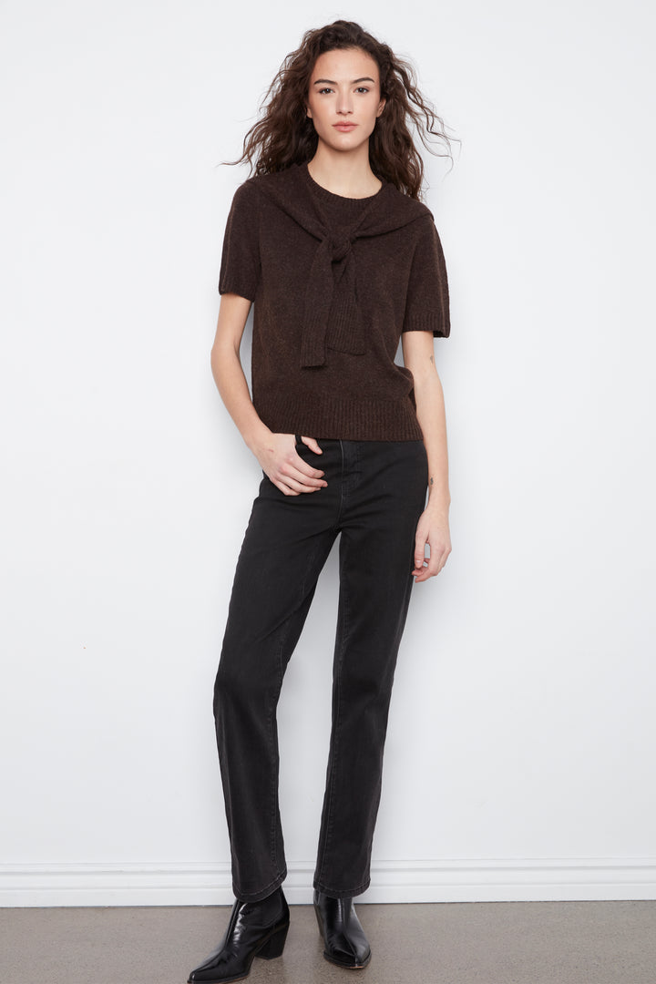 A woman modeling a dark brown short-sleeve knit top with a large front tie detail, paired with black pants and black ankle boots, standing against a white wall.
