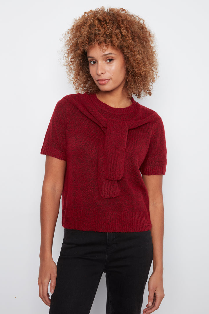 A woman with curly light brown hair wearing a short-sleeve burgundy knit sweater with a knotted collar and black pants, standing against a plain white background.