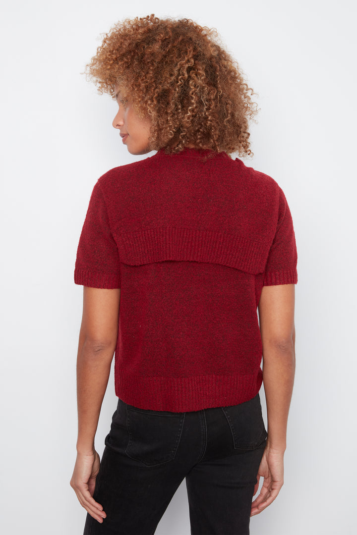 A woman with curly brown hair is shown from the back wearing a short-sleeve knit sweater in a deep burgundy red, paired with black pants, against a plain white studio background.