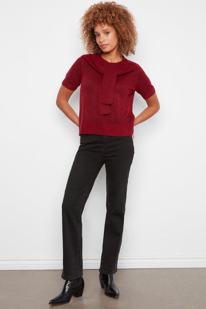 A model with curly brown hair stands against a white wall wearing a short-sleeve burgundy knit top with a large folded collar detail, black pants, and black ankle boots.