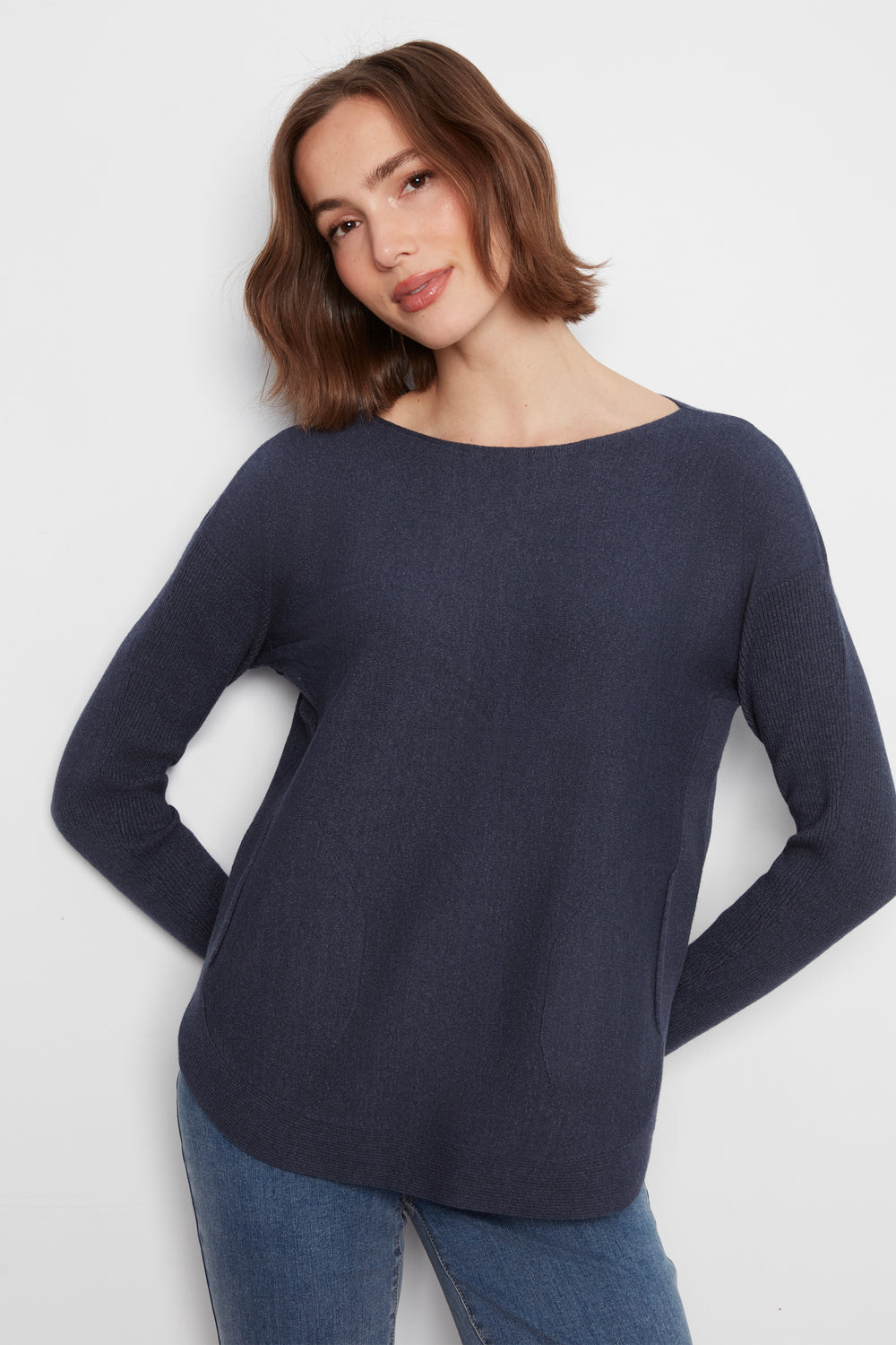 A woman wearing a dark navy blue long-sleeve knit top with a wide boat neckline, paired with blue jeans, against a plain light gray background.